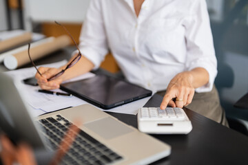 A female architect is focused on her laptop while working on design plans, surrounded by various architectural materials and blueprints in her modern office