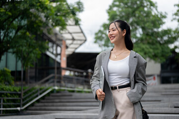 An attractive Asian businesswoman stands in the city with a laptop in her hand, looking away.