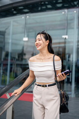 An attractive Asian woman with a smartphone in her hand is walking out of the shopping mall.