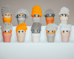 Colourful plastic eggs with wobbly eyes and knitted hats, aligned horizontally and sitting in  bamboo egg holders against a grey background. 