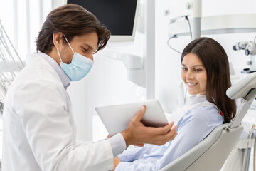 Fototapeta premium Dentist doctor in medical mask showing patient results of treatment on digital tablet, dental office interior