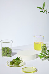 In the center is an empty pedestal in round shape for cosmetic product made from olive oil, surrounded by plenty glassware containing fresh olive and yellow olive oil. Photo from high angle shot