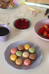 Plate of pastel macarons, cookies and chocolate, cup of tea of coffee, glass of bubble water, various berries, books and accessories on the table. Selective focus, pastel colors.