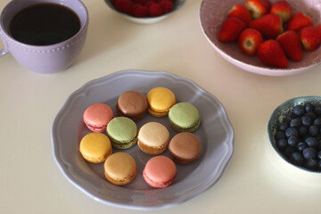 Plate of pastel macarons, cookies and chocolate, cup of tea of coffee, glass of bubble water, various berries, books and accessories on the table. Selective focus, pastel colors.