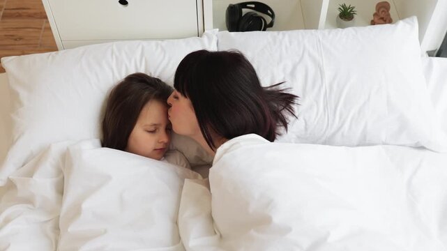 Mother showing affection by kissing her sleeping child in bed during the morning. Image represents love, comfort, family bond, and warmth in a cozy home environment.