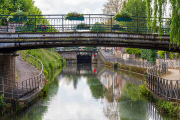 Historic iron bridge over a scenic canal in Montargis, France, with reflections in the calm water and vibrant greenery along the riverside path