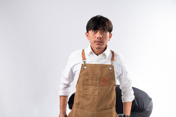 Serious-looking Asian barista wearing a brown apron over a white shirt, standing against a white...