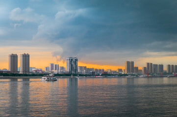 Fototapeta premium Stormy evening sky over the city on the river bank. A pleasure boat with tourists sails along the shore. Red sunset with blue thunderclouds. The Amur River between Russia and China