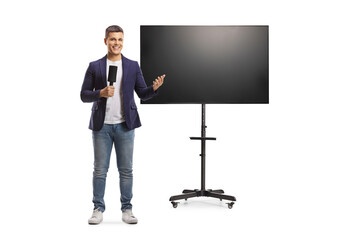 Full length portrait of male presenter holding a microphone and gesturing with hand in front of a tv screen