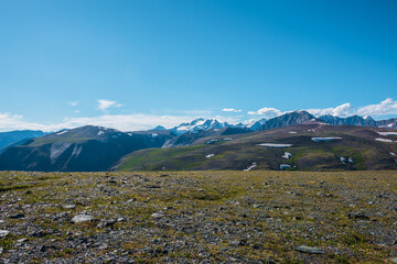 Top view from sunlit grassy stony plain of pass to big snow castle in bright sun under clouds in blue sky. Scenic landscape with green hills and large snow-white rocky mountain far away in sunny day.