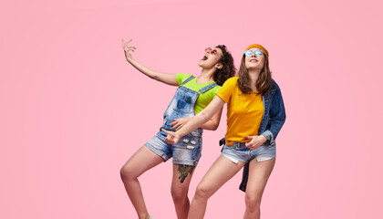 Obraz premium Two young women wearing casual outfits joyfully dancing against a pink background, expressing fun and happiness.