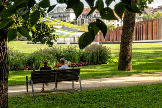 A couple relaxes on a wooden bench enjoying a sunny day in a lush green park. Quiet moments like this highlight the vital role of urban green spaces for relaxation and everyday community wellness.