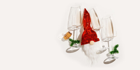 Minimal New Year party concept, flat lay white wine in champagne glasses, Christmas green tinsel, shiny red gnome toy for bottle decor, xmas decor. Top view wineglass sunlight, banner, copyspace
