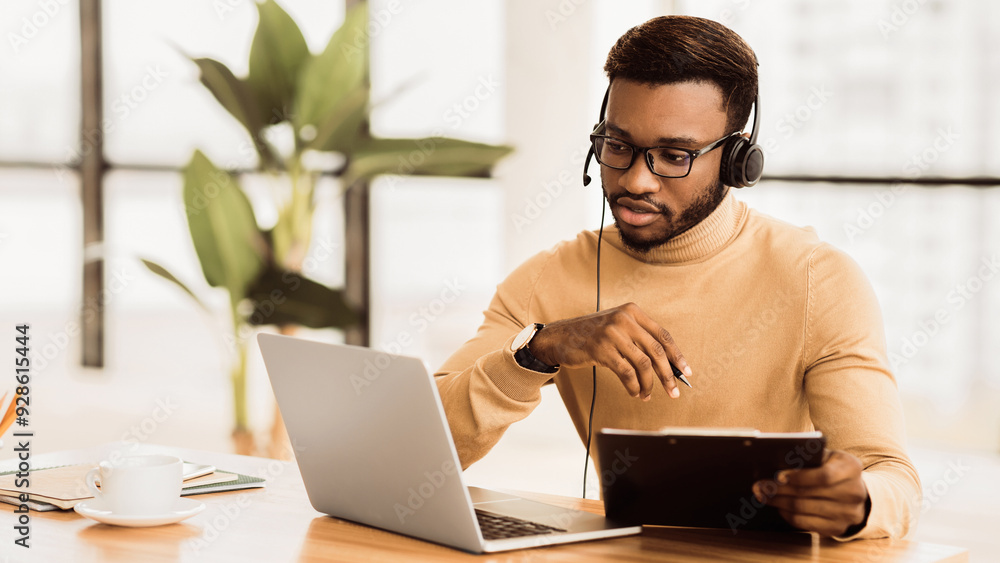 Poster serious african american support operator with headset looking at laptop, writing documentation. emp
