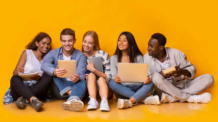 Group of multiracial students studying for university exams, sitting on floor, using laptop, digital tablet, holding books, yellow background