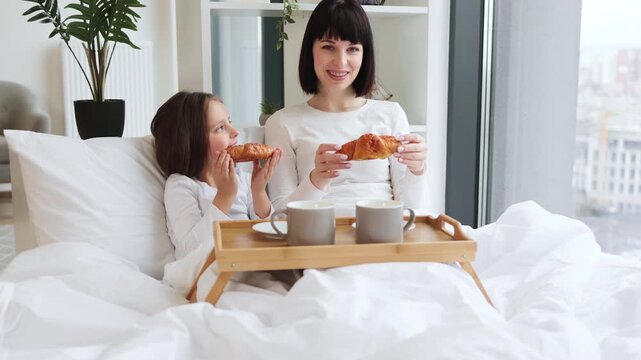 Mother and daughter enjoying breakfast in bed together. They are sharing croissants and drinks, creating a cozy and loving atmosphere. Bright and intimate indoor setting.