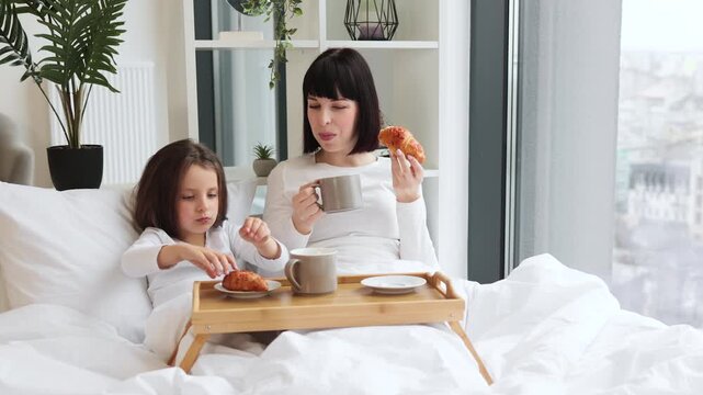 Mother and daughter having breakfast in bed, enjoying croissants and hot drinks. Bright morning light, cozy room with plants, bonding moment between parent and child, relaxation, and comfort.