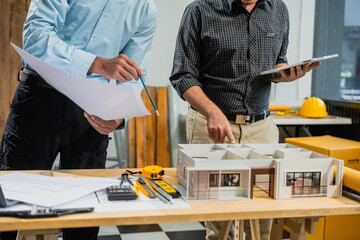 A Caucasian middle-aged male engineer contractor and an Italian architect engineer are seated at a desk, discussing renovation plans with blueprints and a house model in front of them.