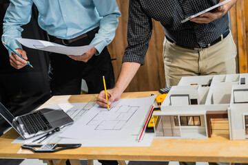 A Caucasian middle-aged male engineer contractor and an Italian architect engineer are seated at a desk, discussing renovation plans with blueprints and a house model in front of them.