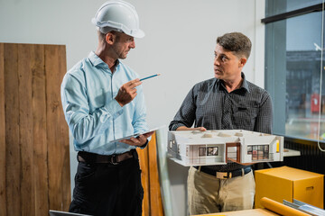 A Caucasian middle-aged male engineer contractor and an Italian architect engineer are seated at a desk, discussing renovation plans with blueprints and a house model in front of them.