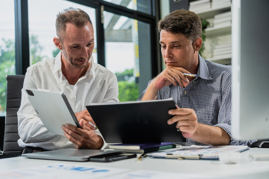 Caucasian middle-aged male businessperson and an Italian accountant are seated at a desk, engaging in a professional meeting, discussing various aspects of technology and innovation in their fields.
