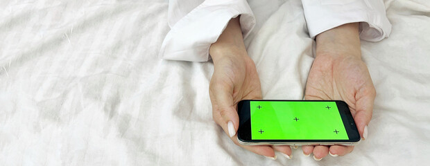 Female hands in a white shirt against the background of a bed hold a phone with a green screen. Mobile Phone Day. Mobile Phone Give Up Day