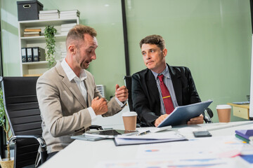 Caucasian middle-aged male businessperson and an Italian accountant are seated at a desk, engaging in a professional meeting, discussing various aspects of technology and innovation in their fields.