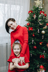 a 30-year-old woman in a red dress hugs her 7-8 year old daughter near the Christmas tree at home in the living room, photo with noise effect