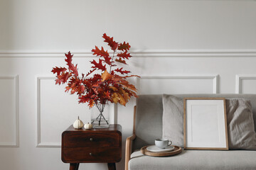 Red oak tree leaves, branches in vase on wooden night table. Linen midcentury sofa with cup of...