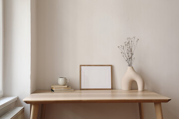 Horizontal empty wooden picture frame mockup. Boho shaped vase, dry flowers on table. Cup of coffee, old books. Working space, home office. Art, poster display. Modern interior. Beige wall background.