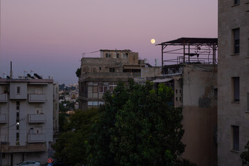 Fototapeta premium Full moon over aging buildings in South Tel Aviv, showcasing a raw urban landscape with visible antennas, cables, and weathered facades under a twilight sky.