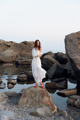 serene woman in white dress and red shoes standing on ocean rock