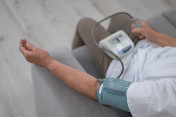 Mature Caucasian woman measuring blood pressure while sitting on sofa at home. 