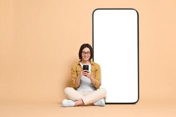 Beautiful young woman with smartphone sitting near big mobile phone with blank screen on dark beige...