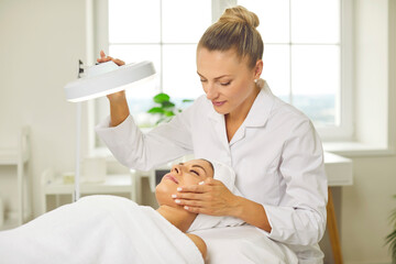 Young professional woman cosmetologist doing skin care facial treatment examining its condition for female client with lamp in beauty clinic. Cosmetology and beauty procedure concept.