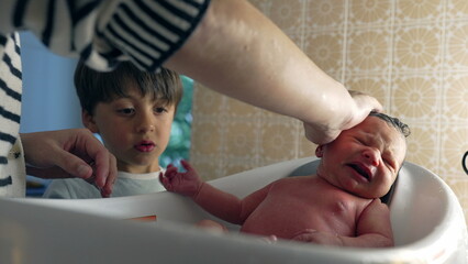 Mother bathing a newborn in a small tub with an older sibling observing, family bonding and care....