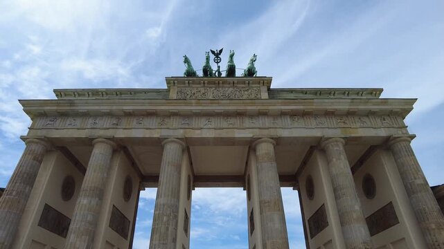 Perspective view from below the Brandenburg Gate. The horses at the top of the gate stand out against a blue sky dotted with subtle, striking clouds. camera tilt up movement 