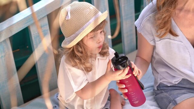 Little blonde curly-haired girl drinks water from plastik bottle that her mother gives her on hot sunny day, outdoors in tropical place. On her head is hat to protect from sun