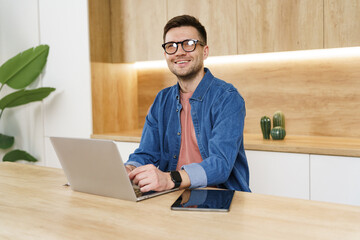 A man in casual attire, working with a laptop in a modern, minimalist office, smiling confidently.