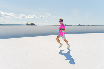 A man running in a bright pink athletic shirt on a wide, open path near the waterfront, under a clear, sunny sky.