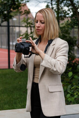 A blonde woman in a light blazer is standing outdoors, focusing on adjusting the settings of her DSLR camera. 