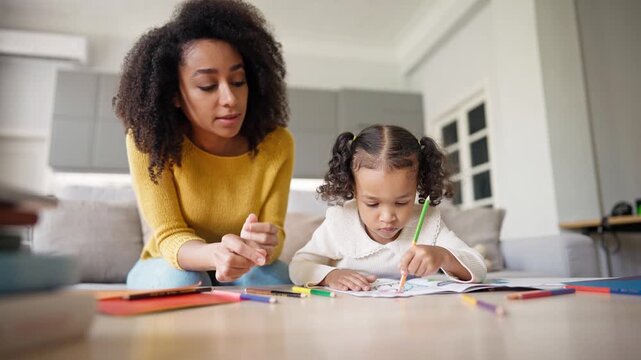 African American mother teaching her loving daughter drawing picture. Obedient girl colouring lines with pencils on colouring page. Mom looking at creative process. Recreation concept.