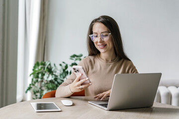 A woman with glasses smiles while working at a wooden table, checking her phone with a laptop and tablet nearby.