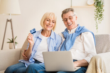 Obraz premium Excited older couple doing online shopping on laptop, woman holding credit card, sitting on sofa at home