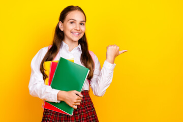 Photo portrait of charming teenager pupil girl hold copybooks point empty space wear trendy uniform isolated on yellow color background