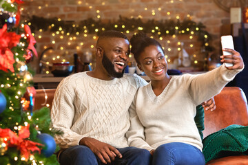 Fototapeta premium Festive selfie. Joyful african american family taking self-portrait, sitting in decorated living room, celebrating Christmas together