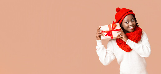 Interested black woman in red hat and scarf checking on her christmas gift over beige background, copy space