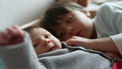 Older sibling lies beside a newborn baby, gazing affectionately, capturing a sweet and tender...