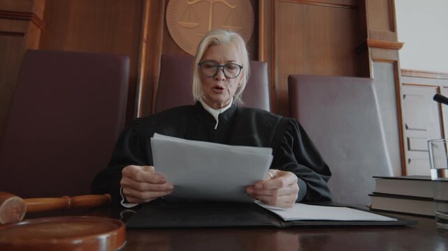 Caucasian mature blond judge hitting sound block with wooden gavel, putting on her glasses and reading out indictment in courtroom
