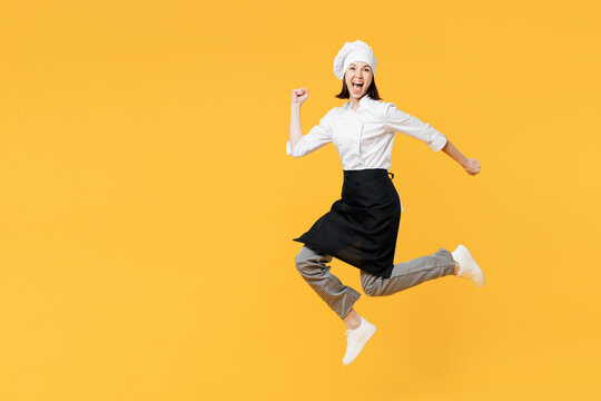 Full body side profile view fun young chef cook baker woman wear white shirt black apron uniform toque hat jump high run fast hurry up isolated on plain yellow background studio. Cooking food concept.